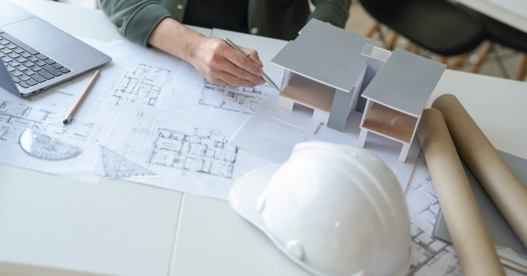 TGA-Planungsleistungen Ein Bauplan mit einem Laptop, einem Helm und Modellen auf einem Tisch.